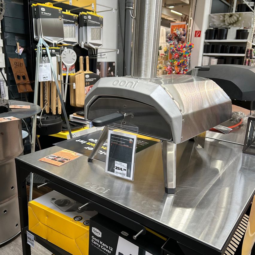 Ein silberner tragbarer Pizzaofen ist in einem Geschäft ausgestellt und hat einen Preisschild. Im Hintergrund befinden sich verschiedene Küchenutensilien und Artikel.