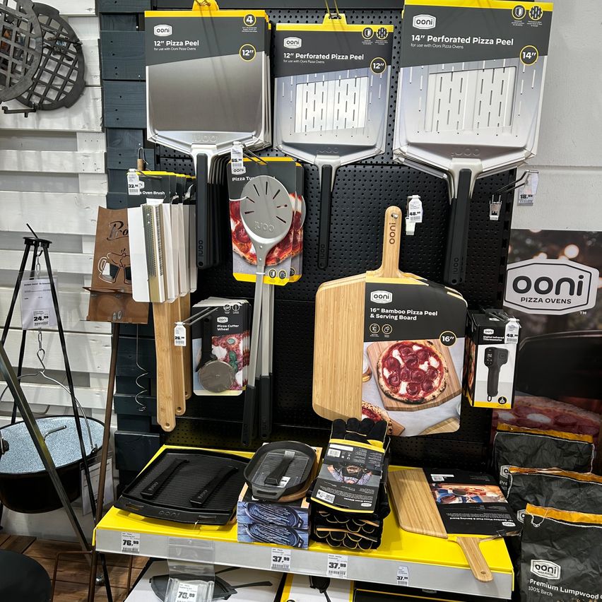 Eine Ausstellung von Ooni-Pizzenschaufeln, Ofenhandschuhen und Pizzaschneidern. Verschiedene Größen von Pizzaschaufeln hängen an einer Lochwand, und einige sind auf gelben Tabletts. Es gibt auch Pizzaöfen und Zubehör auf dem Boden.