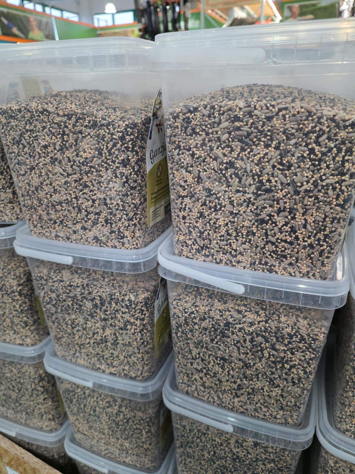 Mehrere durchsichtige Plastikbehälter mit Vogelfutter, die aufeinandergestapelt sind. Jeder Behälter hat ein Etikett, das Ganza liest.