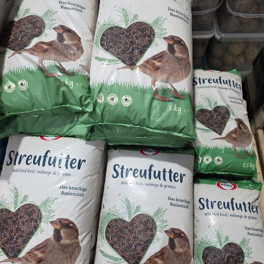 Gestapelte Säcke mit Wildvogelfutter, gekennzeichnet mit 'Streufutter', zeigen einen Vogel auf der Vorderseite mit dem Text 'Das knackige Basismüsli'. Jeder Sack hat ein anderes Gewicht, von 2,5 kg bis 5 kg.