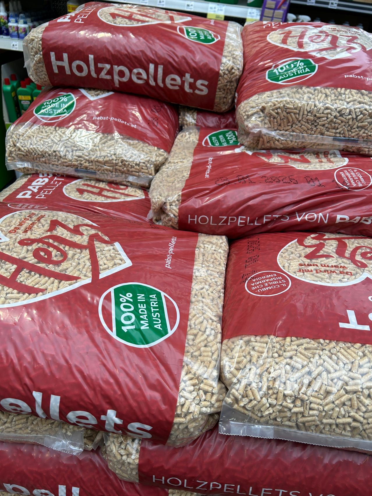 Mehrere Säcke mit Holzpellets sind gestapelt. Jeder Sack hat ein Etikett mit Text und Logos. Ein Sack ist mit '100% MADE IN AUSTRIA' gekennzeichnet. Die Säcke sind rot und haben verschiedene Logos und Texte auf ihnen.