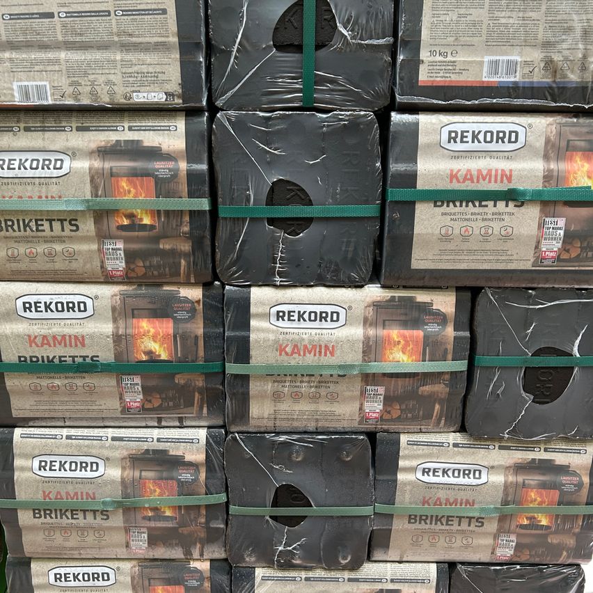 Gestapelte Schachteln von Rekord Kamin Briketts, jede mit einem Bild eines Feuers in einem Ofen, umwickelt mit grünem Klebeband. Die Schachteln sind ordentlich aufeinandergestapelt.