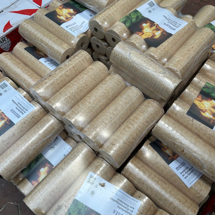 Mehrere Bündel komprimierter Holzpellets, wahrscheinlich für den Einsatz in Kaminen oder Öfen, sind auf einem Boden gestapelt. Die Pellets sind in Plastik verpackt, und jedes Bündel hat ein Etikett mit einem Feuerbild.