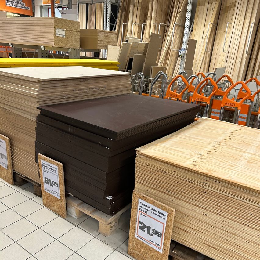 Ein Stapel brauner Sperrholzplatten zum Preis von 81,99, neben einem Stapel Holzbrettern zum Preis von 21,99. Die Platten sind auf einem weißen Fliesenboden angeordnet.