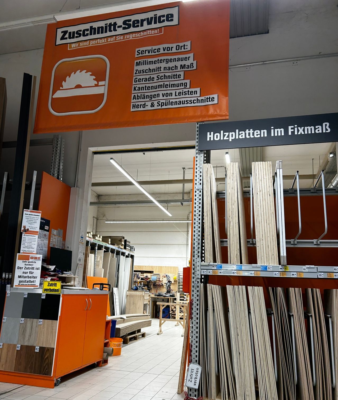 Eine Innenansicht eines Baumarkts zeigt ein Regal mit Holzbrettern, die mit Holzplatten im Fixmaß beschriftet sind. Ein Schild darüber lautet Zuschnitt auf Sie zugeschnitten! mit verschiedenen Dienstleistungen aufgelistet.
