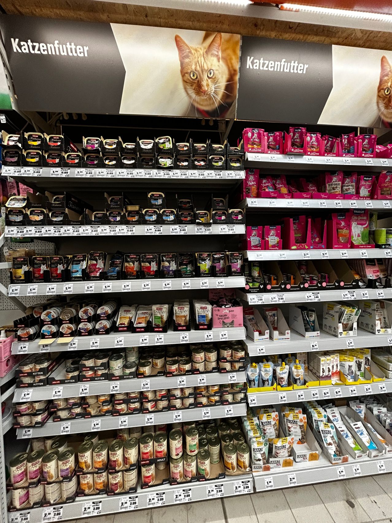 Ein Supermarktregal mit verschiedenen Katzenfutterprodukten, einschließlich Dosen, Beuteln und Tüten, mit einem Katzenposter im Hintergrund.