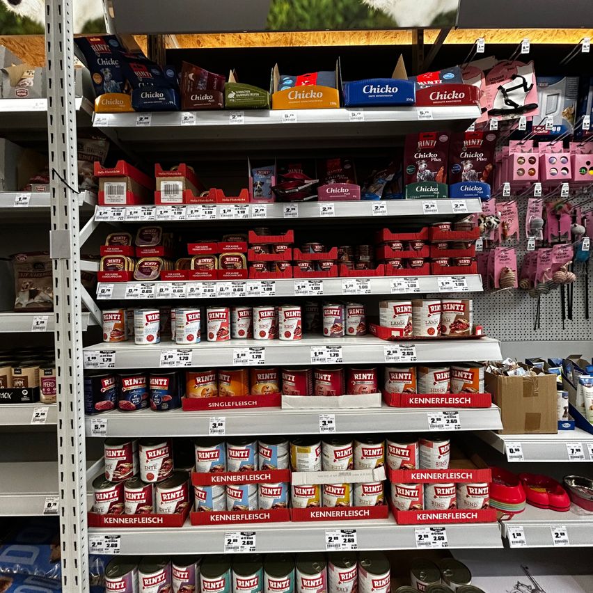Ein Supermarktregal mit Regalen voller Tierfutterprodukte, einschließlich Dosen, Beuteln und Tüten, wobei die Marke 'Rinti' prominent hervorgehoben wird. Verschiedene Größen und Geschmacksrichtungen sind zu sehen, mit sichtbaren Preisschildern.