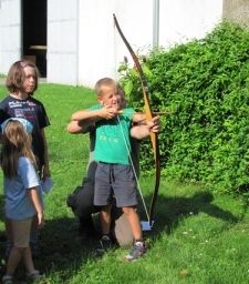 Ein Junge zielt mit einem Bogen, während er in einem Grasfeld steht, drei Kinder und eine Person beobachten ihn. Im Hintergrund ist ein weißes Gebäude mit einer offenen Tür zu sehen.