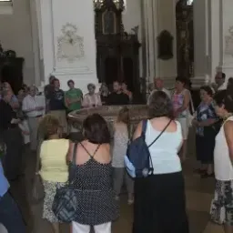 Eine Gruppe von Menschen ist in einer Kirche versammelt, diskutiert und hört einem Sprecher zu. Einige tragen Rucksäcke, und in der Mitte befindet sich ein runder Tisch mit einem Taufbecken.