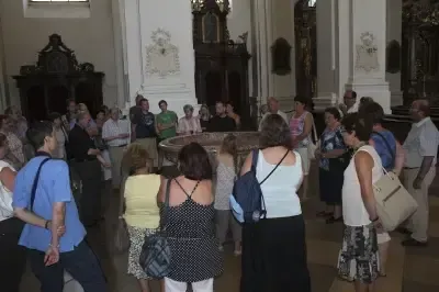 Eine Gruppe von Menschen ist in einer Kirche versammelt, diskutiert und hört einem Sprecher zu. Einige tragen Rucksäcke, und in der Mitte befindet sich ein runder Tisch mit einem Taufbecken.