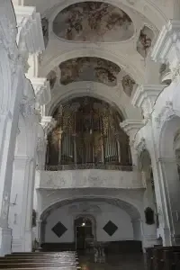 Das Innere einer Kirche mit einer großen Orgel über dem Eingang, kunstvollen Deckenverzierungen und einer langen Reihe von Kirchenbänken.