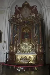 Ein reich verzierter Altar mit goldenen Akzenten und Statuen steht in einer Kirche. Eine große Uhr ist darüber angebracht. Ein roter Seilzaun umgibt den Altar, und ein Blumenstrauß liegt auf dem Boden.
