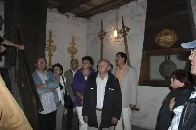 Eine Gruppe von Menschen steht in einem kleinen Raum mit weißen Wänden und beobachtet eine Ausstellung. Der Raum hat Holzbalken und eine an der Decke hängende Lampe.