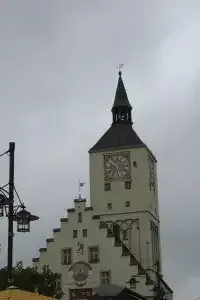 Ein hoher Uhrturm steht hervorgehoben, mit einer Uhr und einem Kirchturm. Der Turm ist Teil eines größeren Gebäudes, und es gibt mehrere Fenster. Eine Flagge weht auf dem Turm.