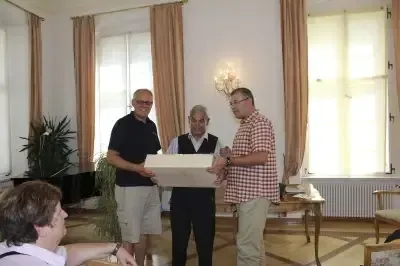 Drei Männer halten eine Kiste, während ein vierter Person beobachtet. Sie sind drinnen, in der Nähe einer Pflanze und eines Klaviers. Fenster mit Vorhängen befinden sich hinter ihnen.