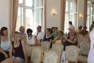 Eine Gruppe von Menschen sitzt in einem Raum mit Stühlen und Fenstern. Einige machen Fotos, während andere zur Kamera schauen. Eine Person steht an der Seite, und es gibt Lichter an der Wand.