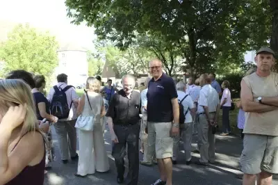Eine Gruppe von Menschen steht im Freien in einem Park mit Bäumen und Gebäuden im Hintergrund. Einige tragen Uhren und Brillen, und eine Person hält eine weiße Tasche.