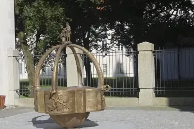 Ein bronzener Brunnen mit kunstvollen Schnitzereien, der eine Statue von zwei Personen zeigt, steht vor einem weißen Gebäude mit einem schwarzen Zaun. Bäume und Schatten sind sichtbar.
