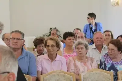 Eine Gruppe älterer Personen mit Brillen sitzt in Stühlen in einem Raum. Einige von ihnen schauen nach vorne, während andere etwas betrachten. Hinter ihnen steht ein Mann und hält eine Kamera.