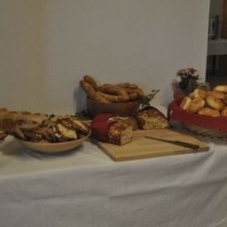 Ein mit einem weißen Tuch bedeckter Tisch zeigt eine Vielzahl von Broten, Gebäck und Bagels. Ein Holzschneidebrett mit Messer, ein Toaster und ein Blumenvase sind ebenfalls vorhanden.