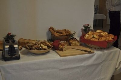 Ein mit einem weißen Tuch bedeckter Tisch zeigt eine Vielzahl von Broten, Gebäck und Bagels. Ein Holzschneidebrett mit Messer, ein Toaster und ein Blumenvase sind ebenfalls vorhanden.