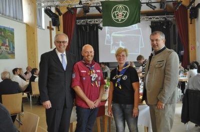 Drei Personen stehen vor einer großen Leinwand mit einem grünen Banner. Sie sind von Menschen umgeben, die an Tischen in einem Saal sitzen.