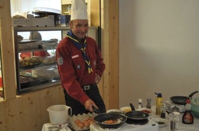 Ein Koch in einem roten Outfit bereitet Essen auf einem Herd in einer Küche mit Vitrine, Holzwänden und verschiedenen Utensilien zu.