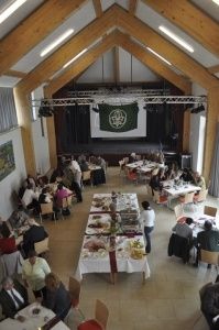 Ein Blick von oben auf einen Speisesaal mit Menschen, die an Tischen sitzen. Der Saal hat Holzbalken, weiße Wände und eine große Leinwand auf der Bühne. Die Tische sind mit weißen Tüchern bedeckt und mit Essen gefüllt.