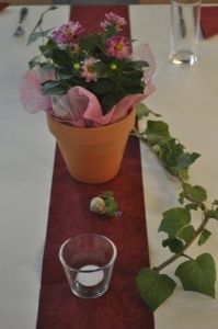 Ein Tisch ist mit einer roten Tischdecke, einem braunen Blumentopf, einer Pflanze mit rosa Blumen, einem Glaskerzenhalter und einem Glasbecher gedeckt. Ein kleines dekoratives Element befindet sich auf dem Tisch.