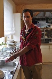 Ein Mann steht in einer Küche und spült Geschirr in einer Spüle ab. Er trägt ein rotes Hemd und eine Schürze mit einem Abzeichen. Auf der Arbeitsplatte stehen Teller, Schüsseln und Gläser. Hinter ihm befindet sich ein Kühlschrank und ein Regal mit verschiedenen Gegenständen.