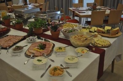 Ein langer Tisch, der mit einer weißen Tischdecke bedeckt ist, ist mit verschiedenen Lebensmitteln gefüllt. Der Tisch ist mit Topfpflanzen und Schüsseln mit Lebensmitteln geschmückt. Es gibt mehrere Stühle und Tische in der Gegend. Es gibt auch einige Edelstahlschalen auf dem Tisch.