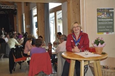 Eine Frau sitzt an einem runden Tisch in einem Essbereich und hält einen Stift. Mehrere Personen sitzen an anderen Tischen, mit einer Blumenvase in der Nähe. Eine Ankündigung befindet sich an einem Aushang.