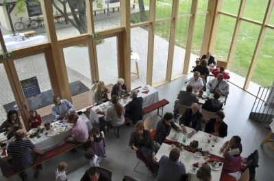 Ein Blick von oben auf einen Essbereich mit mehreren Tischen. Mehrere Personen sitzen an Tischen, einige mit Essen. Der Bereich hat große Fenster und Holzwände. Draußen gibt es eine Rasenfläche und einen Baum.