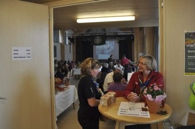 Zwei Frauen stehen an einem Tisch mit einem Blumentopf und unterhalten sich. Es gibt mehrere Menschen, die um Tische herumsitzen und essen. Im Hintergrund ist eine Projektionsleinwand zu sehen. An der Wand befindet sich ein Plakat.