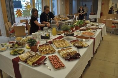 Ein langer Tisch, der mit einer weißen Tischdecke bedeckt ist, ist mit verschiedenen Speisen gefüllt, darunter Desserts, Früchte und andere Gerichte. Zwei Personen stehen hinter dem Tisch, eine bereitet Essen zu. Im Hintergrund stehen mehrere Personen in der Nähe von Tischen.