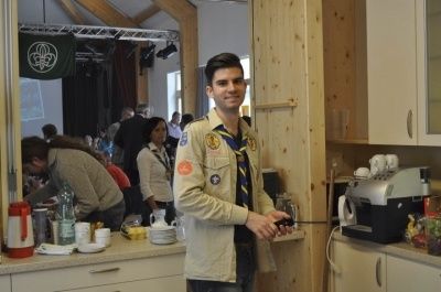 Ein junger Mann in einem Pfadfinder-Uniform steht in einer Küche, hält eine Fernbedienung in der Hand, mit einer Kaffeemaschine und verschiedenen Gegenständen auf dem Tresen hinter ihm.