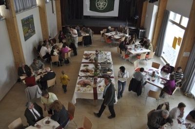 Eine große Gruppe von Menschen isst an einem langen Tisch in einem Speisesaal. Die Tische sind mit Essen bedeckt. Eine Projektionsleinwand befindet sich an der Wand.