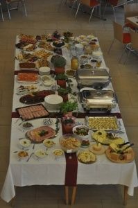 Ein langer Tisch mit einer weißen Tischdecke, bedeckt mit einer Vielzahl von Speisen, einschließlich Fleisch, Gemüse und Desserts. Es sind auch mehrere Serviertabletts und Schüsseln vorhanden.