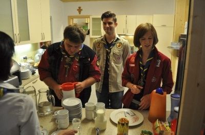 Vier Personen befinden sich in einer Küche. Drei stehen in der Nähe einer Theke mit Tellern, Tassen und Essen. Die vierte Person sitzt ihnen gegenüber.