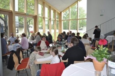 Innenbereich zum Essen mit großen Fenstern. Viele Menschen sitzen an Tischen, einige Kinder anwesend. Topf mit pinken Blumen. Weiße Tischdecken.