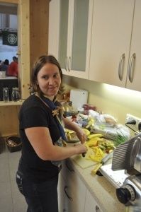 Eine Frau in einem schwarzen Shirt und Schal steht in einer Küche und schneidet Obst und Gemüse auf einem Schneidebrett. Es gibt verschiedene Gegenstände auf der Arbeitsplatte, und Küchenschränke und Geräte sind im Hintergrund.