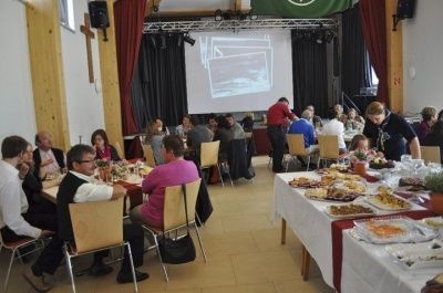 Ein großer Raum mit einer Projektionsleinwand an der Wand, mehrere Personen sitzen an Tischen mit Essen darauf, eine Frau steht und serviert Essen.
