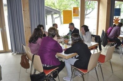 Eine Gruppe von Menschen sitzt an einem Tisch in einem Essbereich und führt ein Gespräch. Der Raum hat Glasfenster und Vorhänge, mit einer Pflanze und Postern an der Wand sichtbar.