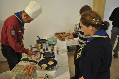 Ein Koch mit Toque bereitet Essen auf einem Herd mit Eiern und anderen Zutaten zu. Eine Frau beobachtet ihn, und ein Mann ist in der Nähe. Verschiedene Lebensmittel und Flaschen stehen auf dem Tisch.