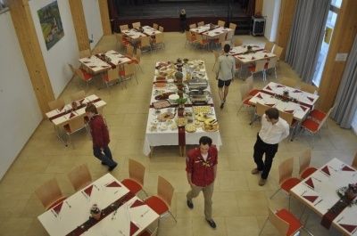 Ein Speisesaal mit mehreren gedeckten Tischen mit Essen, wo mehrere Personen in der Nähe eines langen Tisches mit verschiedenen Gerichten stehen. Der Raum hat eine Bühne im Hintergrund.