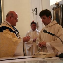 Zwei Priester stehen vor einem Altar, einer erteilt einen Segen, während der andere aufmerksam zuhört. Sie sind von einer kleinen Gemeinde umgeben, mit einem Gemälde an der Wand hinter ihnen.