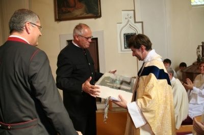 Ein Priester in einer goldenen Robe überreicht einem anderen Mann in einer schwarzen Robe ein Geschenk in einer Kirche. Hinter ihnen klatschen mehrere Menschen. Ein Gemälde hängt an der Wand über ihnen.