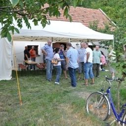 Mehrere Menschen versammeln sich unter einem weißen Zelt in einem Grasgarten mit geparkten Fahrrädern. Ein Haus und Bäume umgeben das Gebiet.
