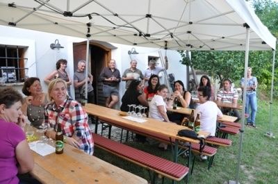 Eine Gruppe von Menschen sitzt an einem langen Holztisch unter einem weißen Zelt. Sie trinken Bier und Wein. Der Tisch ist mit Flaschen, Gläsern und Servietten gefüllt. Sie lächeln und scheinen eine gesellige Zusammenkunft zu genießen.