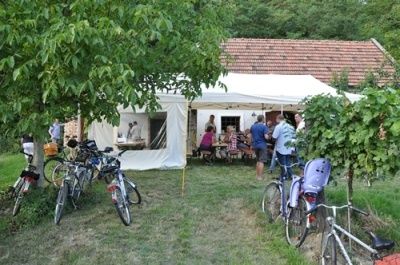 Eine Gruppe von Menschen sitzt und steht um einen Tisch herum unter einem weißen Zelt in einem Hof mit vor dem Zelt geparkten Fahrrädern.